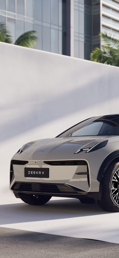 Grey Zeekr X electric SUV seen from the front three-quarter angle, parked beside a white wall with palm trees and glass…