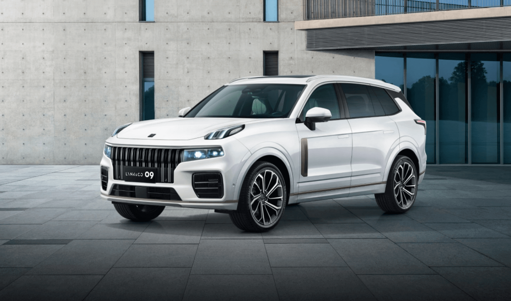White Lynk & Co 09 SUV in three-quarter front view, parked on a stone plaza in front of a concrete building